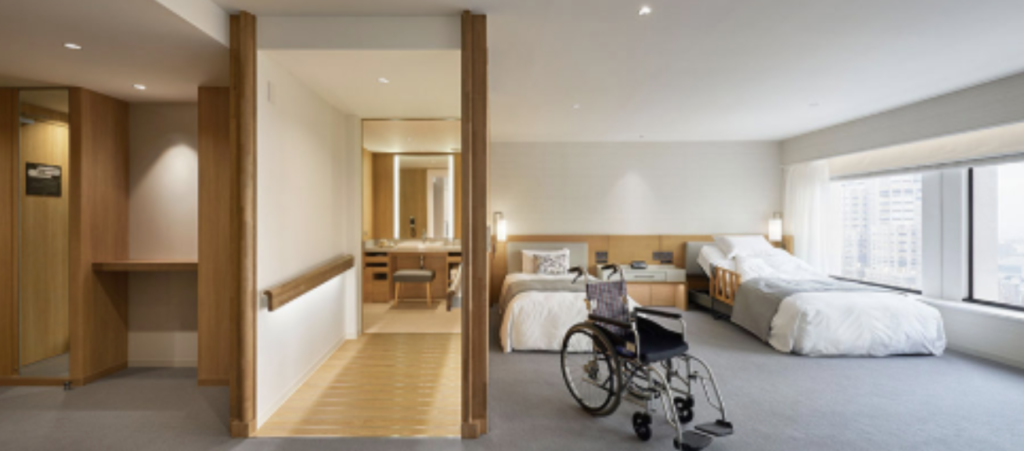 Tekerlekli sandalye erişimine uygun, geniş kapılı ve ferah bir otel odası; odada iki yatak, geniş bir geçiş alanı ve pencereden gün ışığı alan bir iç mekân görülüyor.